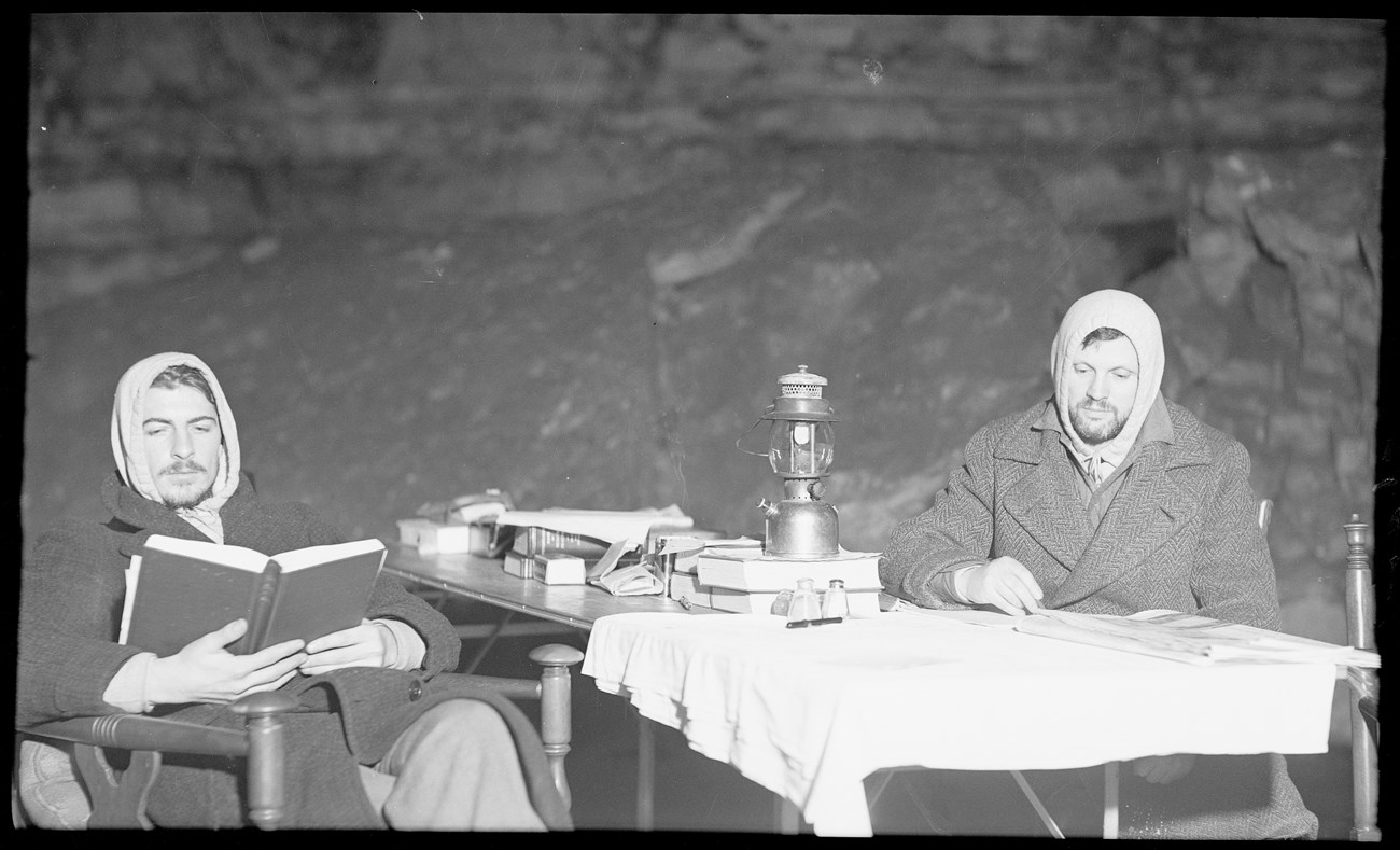 Two men study books and papers while seated at a table with a lantern placed in the middle.