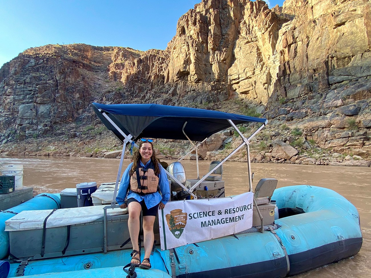 Cavezza leans against a raft with a sign with the NPS logo that says, "Science and Resource Management." Behind here is the Colorado River and the bluffs of the canyon.