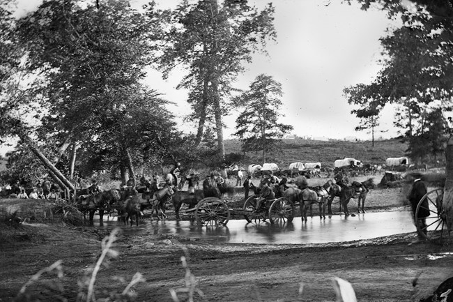 Cedar Mountain horse-drawn artillery crossing a shallow stream in front of a gathering of soldiers and a collection of covered wagons.