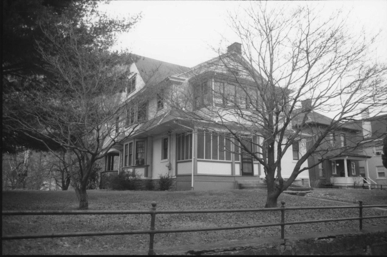 A white and black clapboard house with a gable roof sits on a hill near several trees.