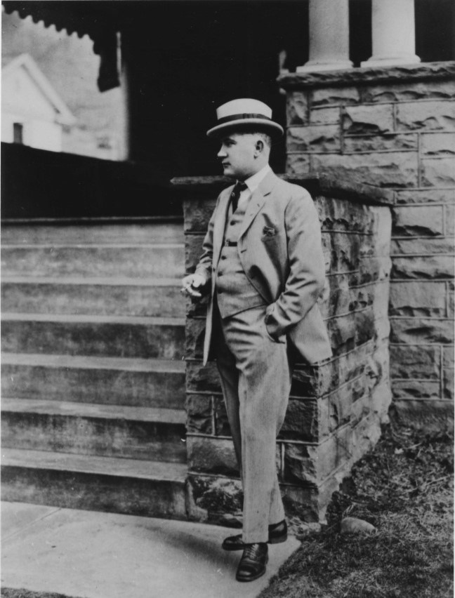 Chafin Photo Portrait A man in a tan suit and hat leans on a stone pedestal near steps.