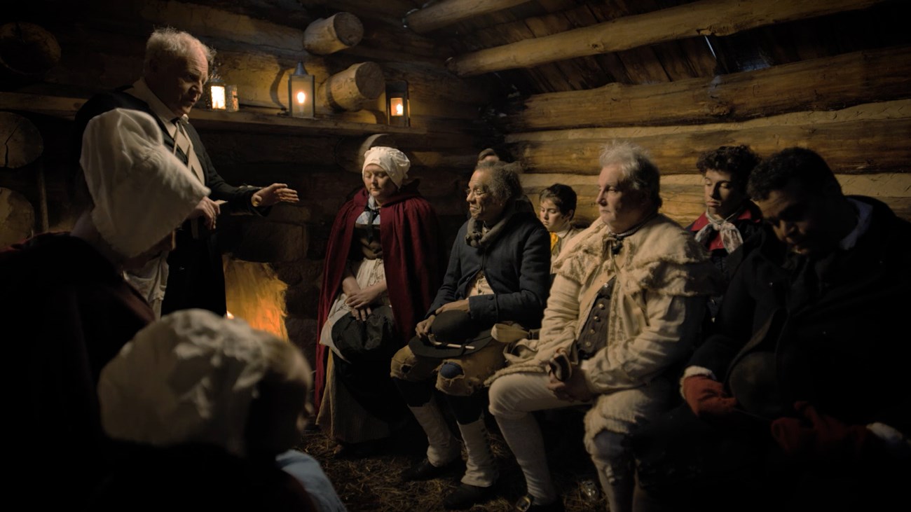a man in a robe speaks to people seated in front of a fire inside a log hut