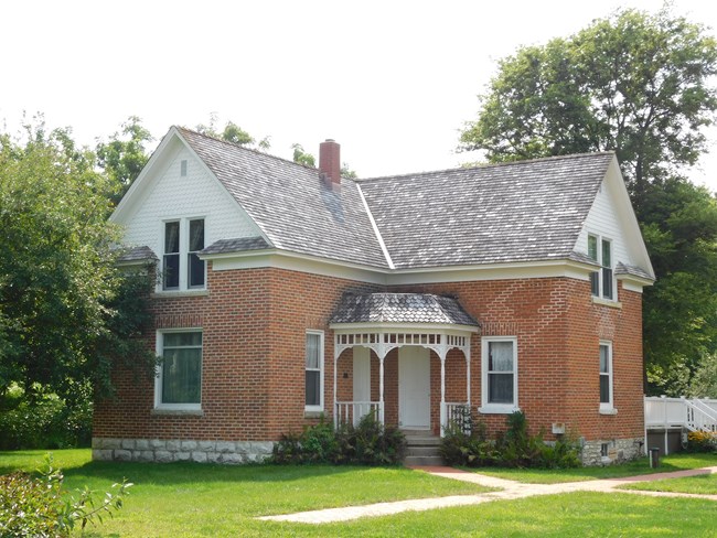 Tan brick, Victorian-era farm house with white scalloped siding near the roofline, gray gabled roof, and stone near the foundation. The house is two stories and sits on an L with a covered entryway at the inner corner