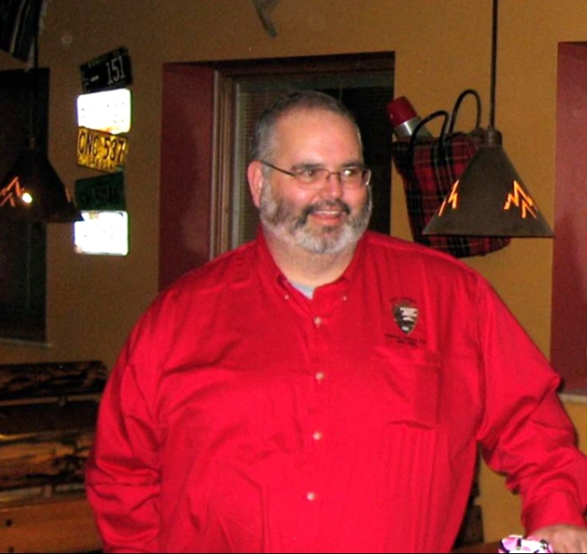 A white man with dark gray hair, white and gray beard, and wearing wire-frame glasses and a bright red button-up long sleeve shirt with the National Park Service arrowhead logo on the front right of it smiles at something off camera in a low-lit room.
