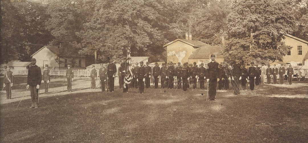 Lieutenant Charles Young, second from left, front of formation, in front of the student military detachment at Wilberforce University,
Courtesy of the Coleman Collection, National Afro-American Museum and Cultural Center, Wilberforce, Ohio. Black and white photo of African American army cadets in uniform standing in formation.