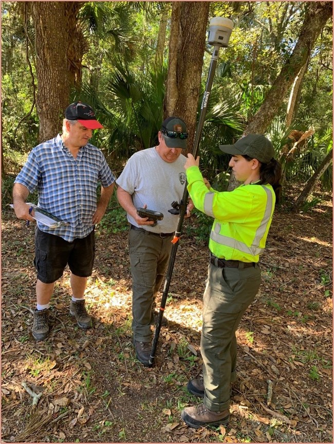 three people standing under trees around a survey pole