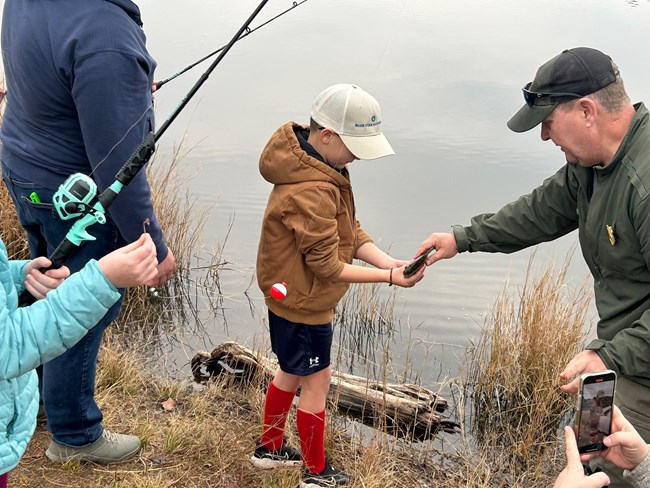 Junior Ranger Angler Fishing Clinic at TIMU Man in uniform hands a fish to a boy in a hat and red sweatshirt