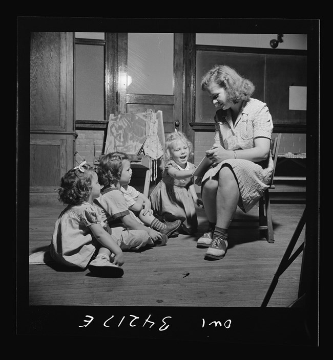 A woman sits on a low chat reading a book to three toddler-age children in a classroom setting