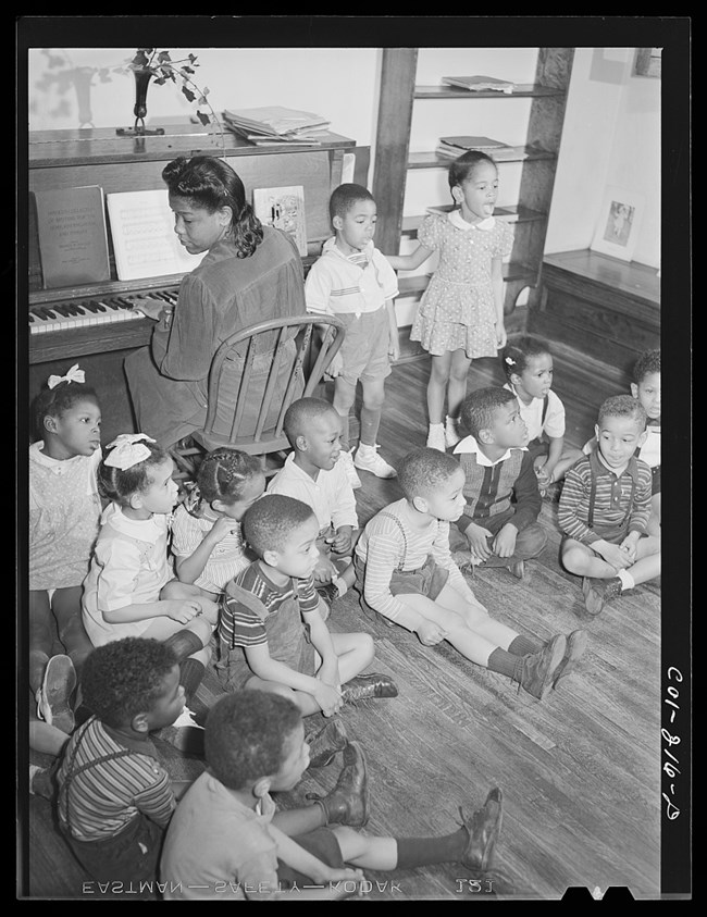 An African American woman sits at a piano while several children sit on the wooden floor behind her