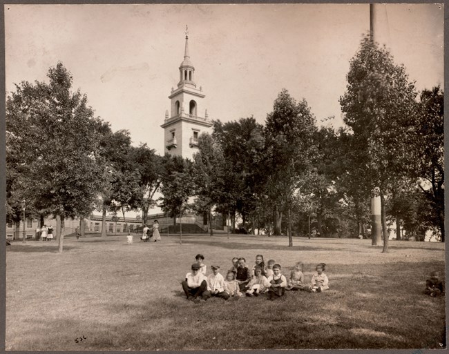 Group of children sitting on grass in a park with the Dorchester Heights Monument in the background.