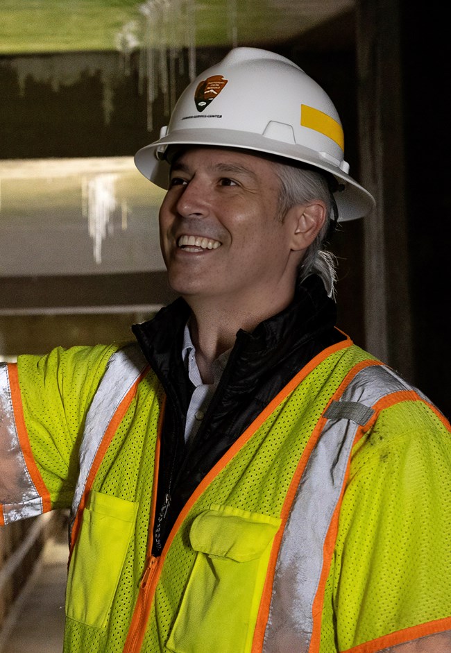 A person in an orange mesh reflective vest and a white hard hat in a lighted boardwalk tunnel.
