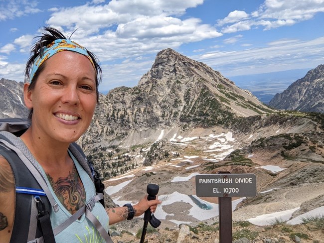 Christina Appleby Woman on a hike smiles while standing near a sign reading Paintbrush Divide, with rugged mountain scenery behind her.