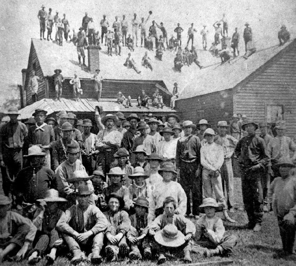 Black and white photo of a large gathering of men sitting on the ground and standing on the roof top of a house in the background.