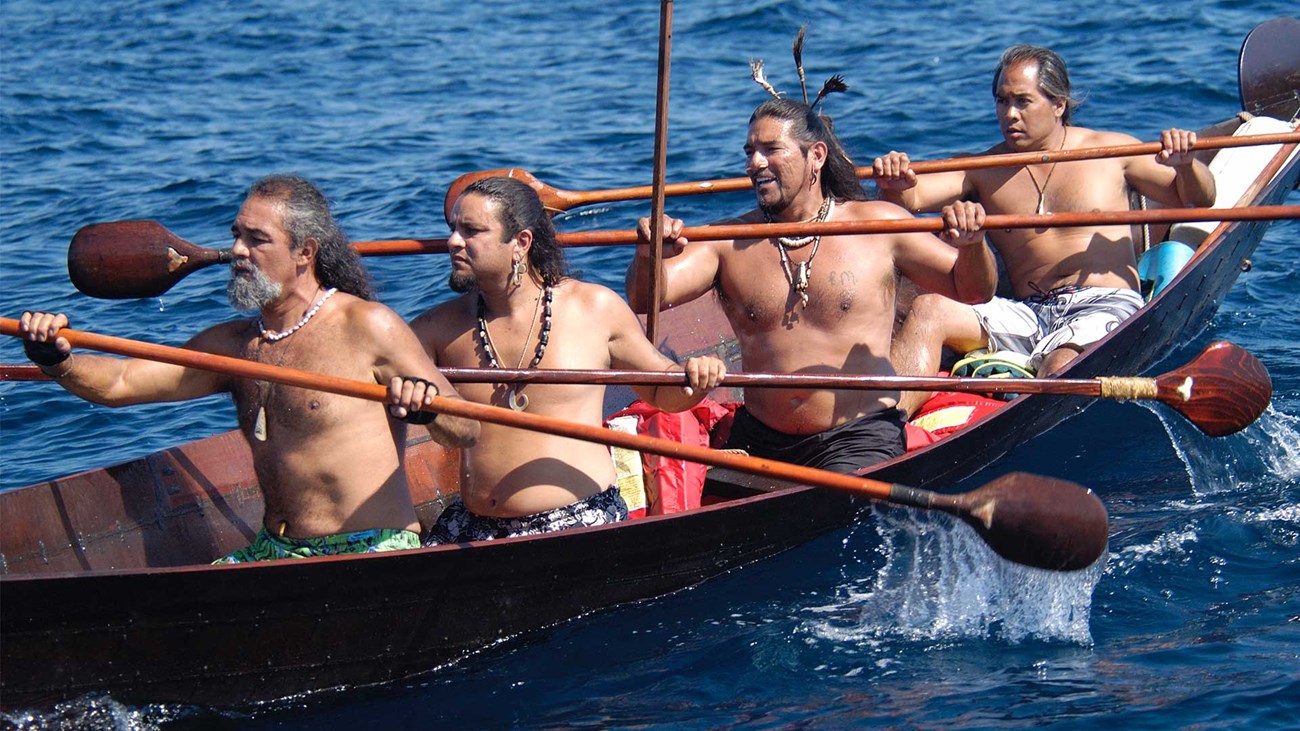 Four topless indigenous men paddle in wooden canoe