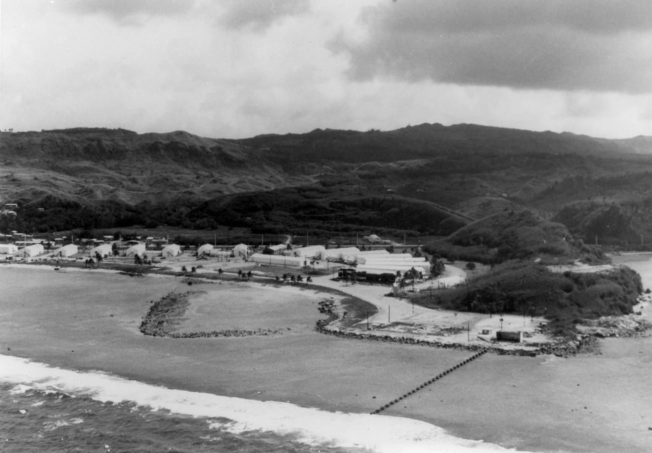 Black and white photo of a series of white buildings built on the flat area next to the ocean. An area has been created offshore for people to swim.