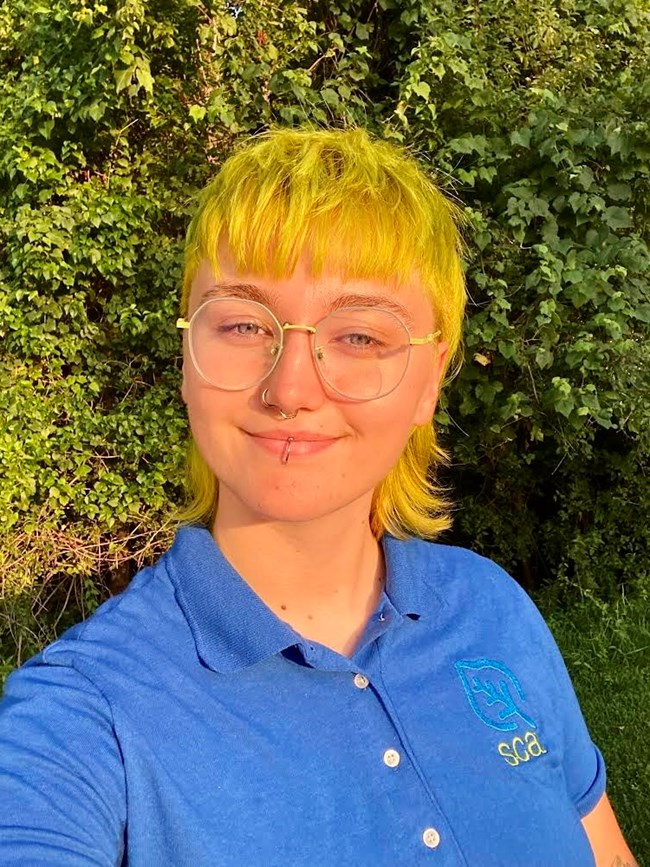 YMCA Partnership camp leader Claire “Twig” Adams A young woman with blonde hair, glasses, and a blue shirt in front of a green bush