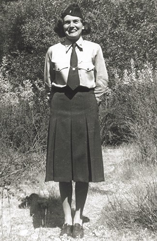 A women wearing a long dark skirt, white blouse, tie and flight cap stands with her hands behind her back smiling at the camera.  Brush can seen behind her.