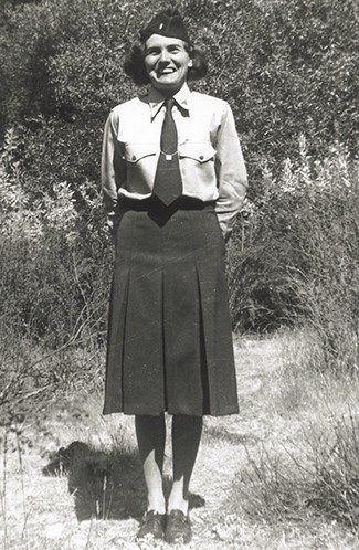 Clara Ann Lausten_1942_Pinnacle NM A women wearing a long dark skirt, white blouse, tie and flight cap stands with her hands behind her back smiling at the camera. Brush can seen behind her.