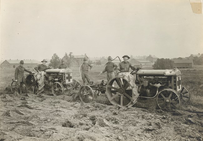 Six white soldiers stand behind and around two tractors in the middle of a field with upturned earth. Two of the men hold scythes. Signs on the tractors display the American Red Cross symbol and read "American Red Cross USA Hospital Fort Des Moines."