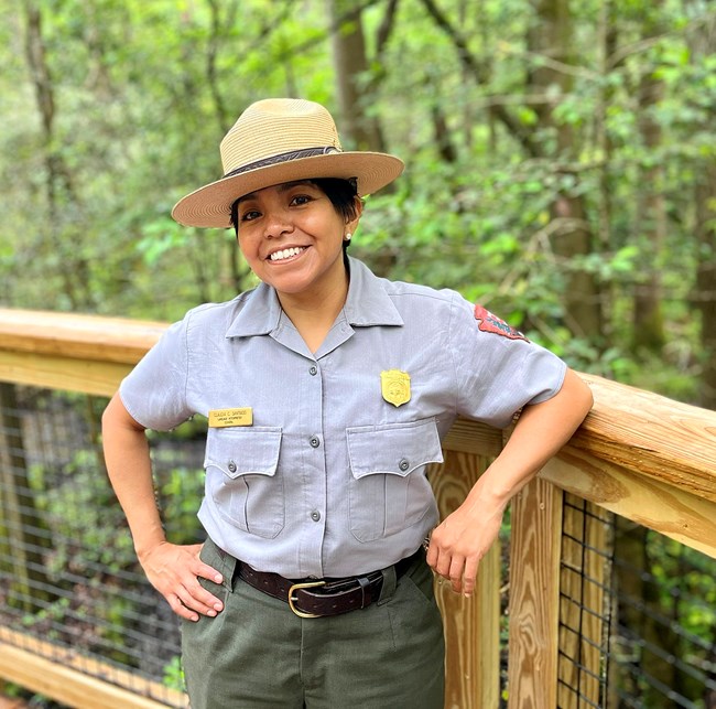 Woman in NPS uniform leans against the railing of a bridge in front of trees with green leaves.
