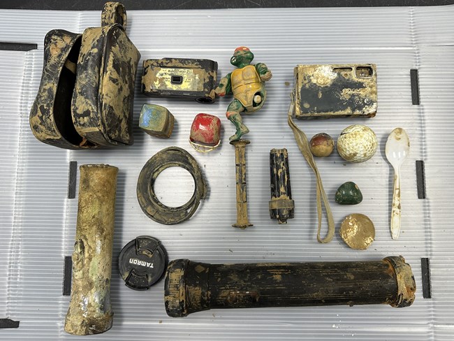 A muddy black camera bag, camera, worn out flashlights, flash cubes, plastic bouncy balls, white golf ball, white plastic spoon and a toy turtle figure laid out on display.