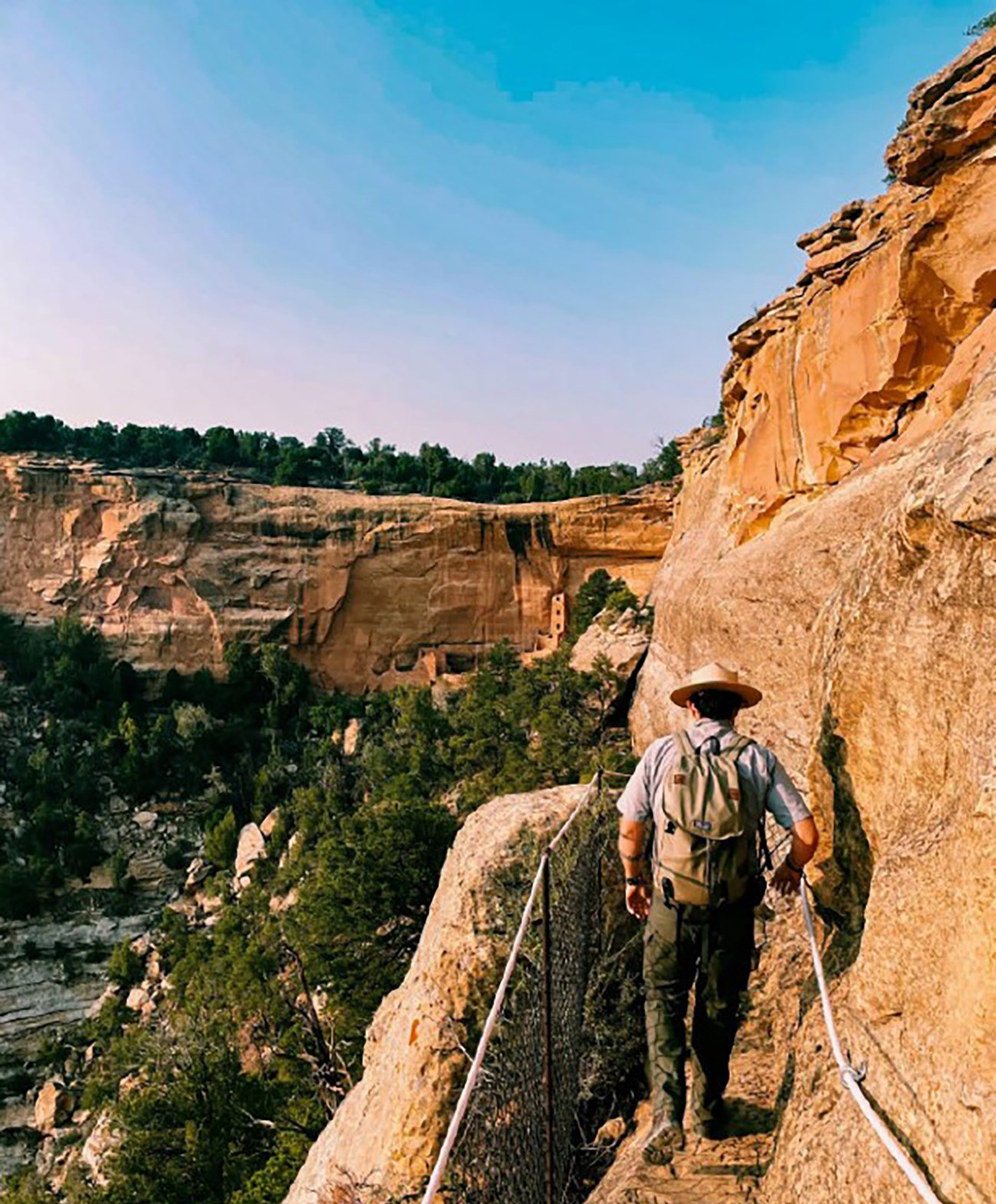 A ranger walks along a narrow trail on a cliffside. Cliff dwellings are visible in the distance.