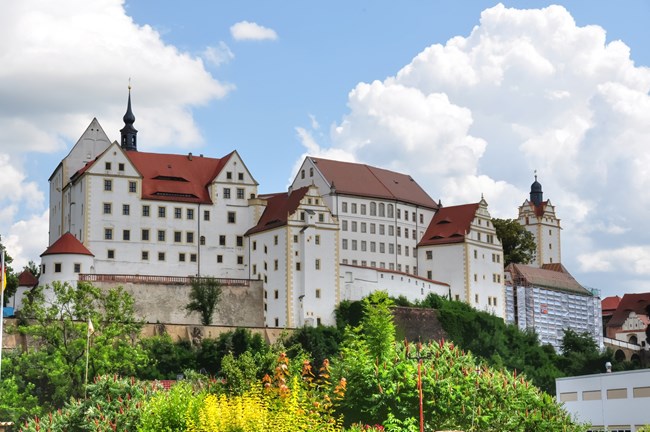Renaissance style castle sitting high on a hill. The castle has multiple buildings and towers with smooth whitewashed walls and a steep red roof. Lush landscaping is visible in the foreground. A partly cloudy sky is visible in the background.