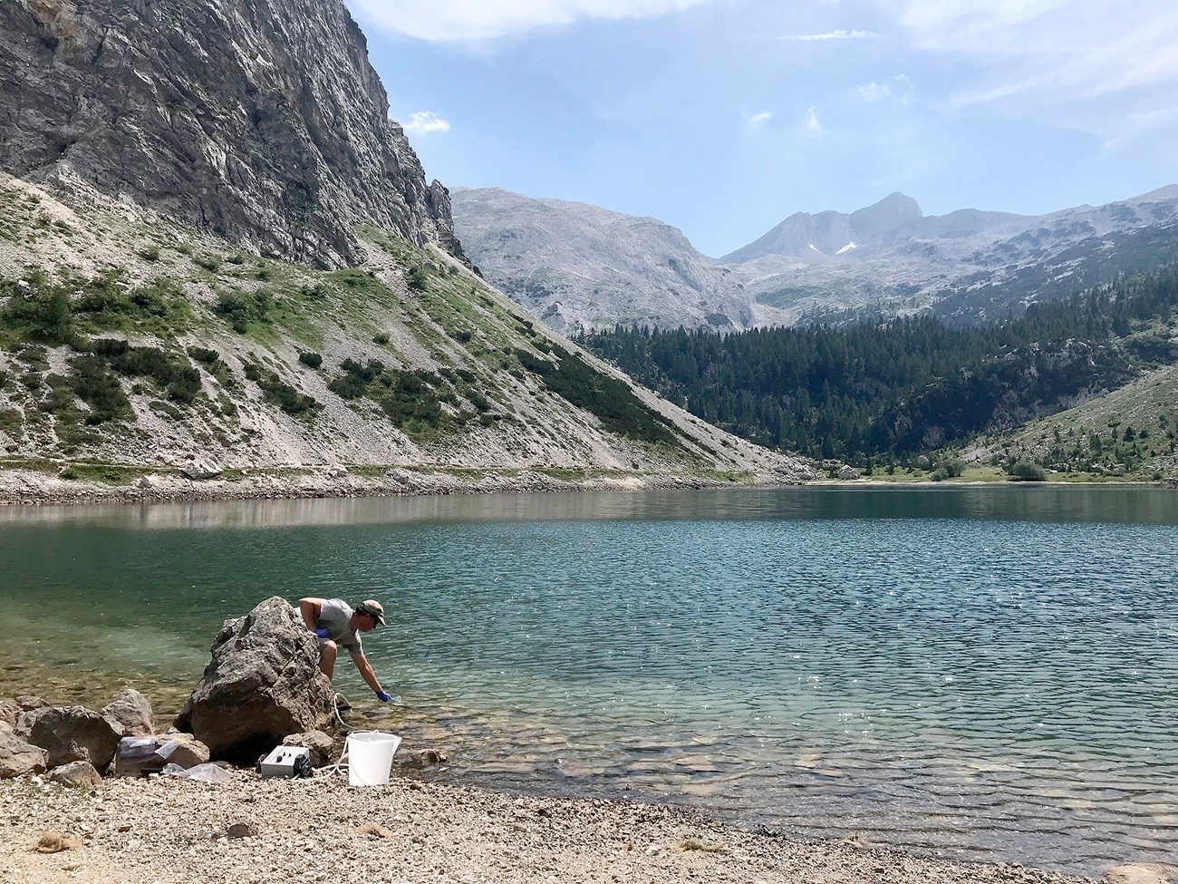 Collecting lake water for eDNA analysis A man in a white shirt and baseball cap dips a sampling cup into a blue lake. he is surrounded by mountains, trees, and blue sky.