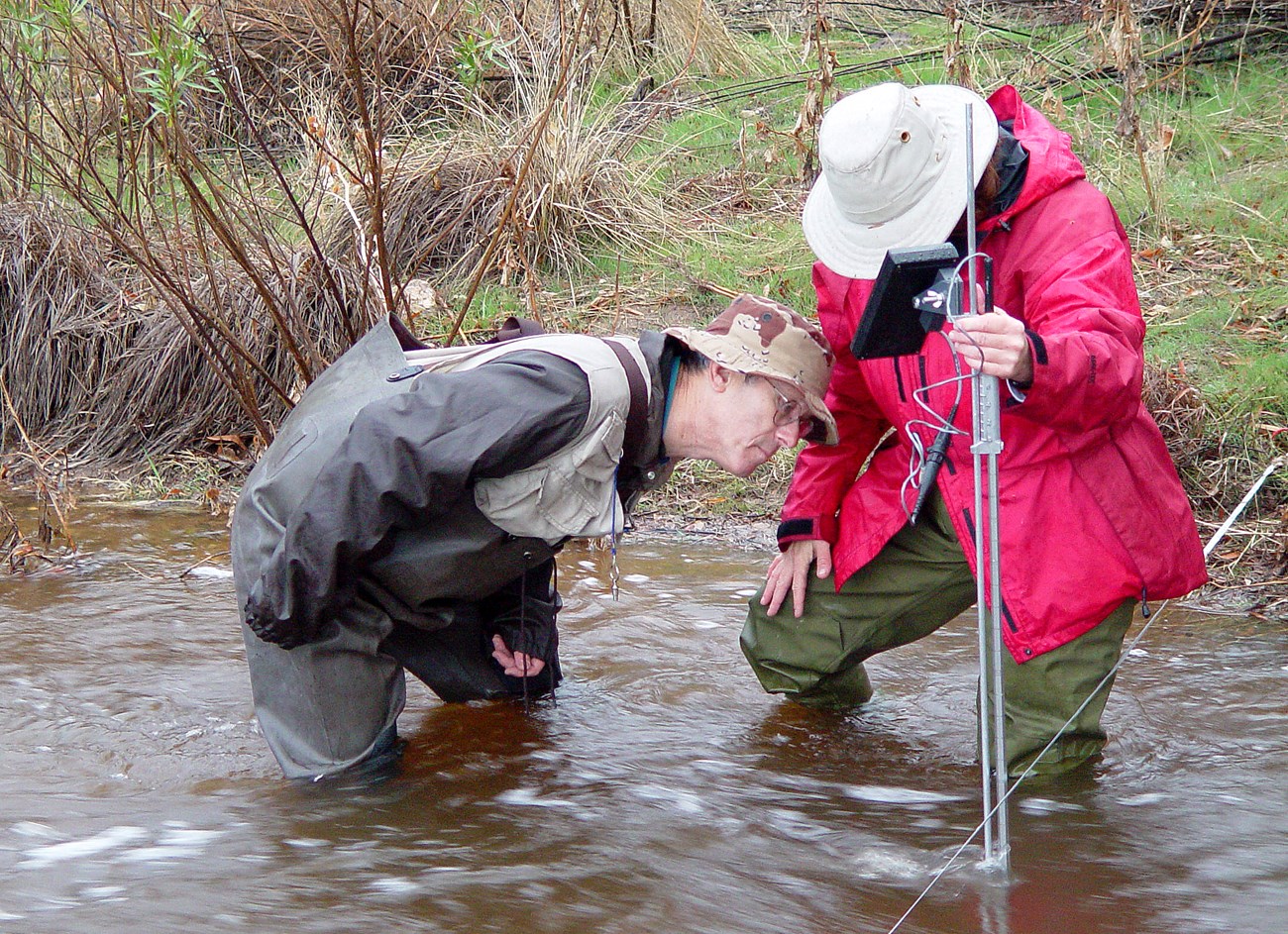 A man bending over to examine an instrument held by a woman. Both are standing in a stream.