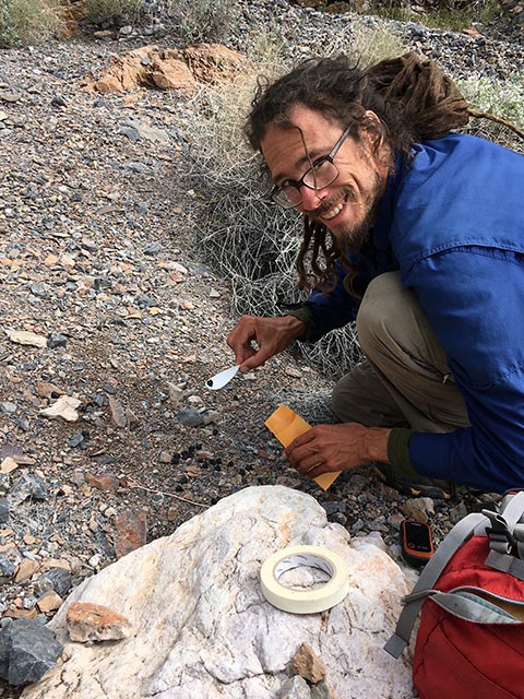 A researcher kneeling down next to a rock with a sliver spoon and envelope collecting small brown scat pellets.