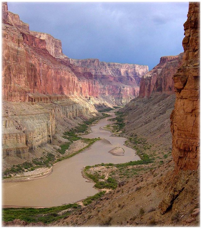 Deep canyon with silty river running through, flanked with narrow  strips of green on both sides. Canyon walls are layers of varying shades of red, tan and brown rock.