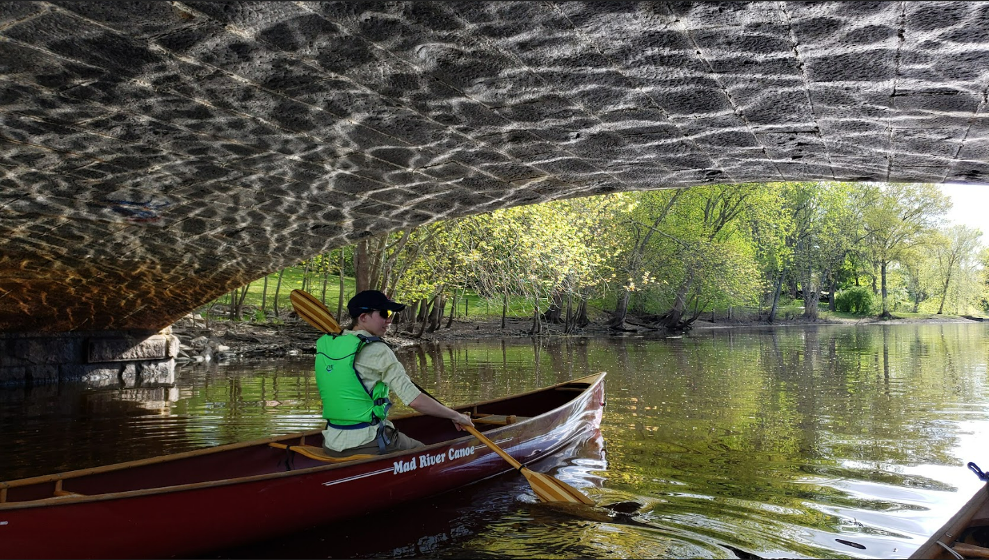 a person paddles a canoe under a bridge that shows the water's reflection in the sun.