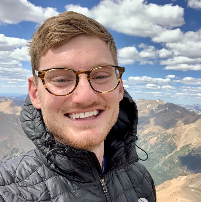 Man smiling atop a mountain with views of more mountains and scattered clouds in background.