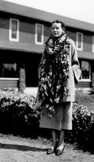 A woman wears flower leis around her neck and long coat.  She stands with her hands in her pockets.