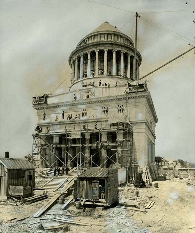 Black and white photo of workers on wood supports on a white tomb, with construction materials scattered in front of the structure