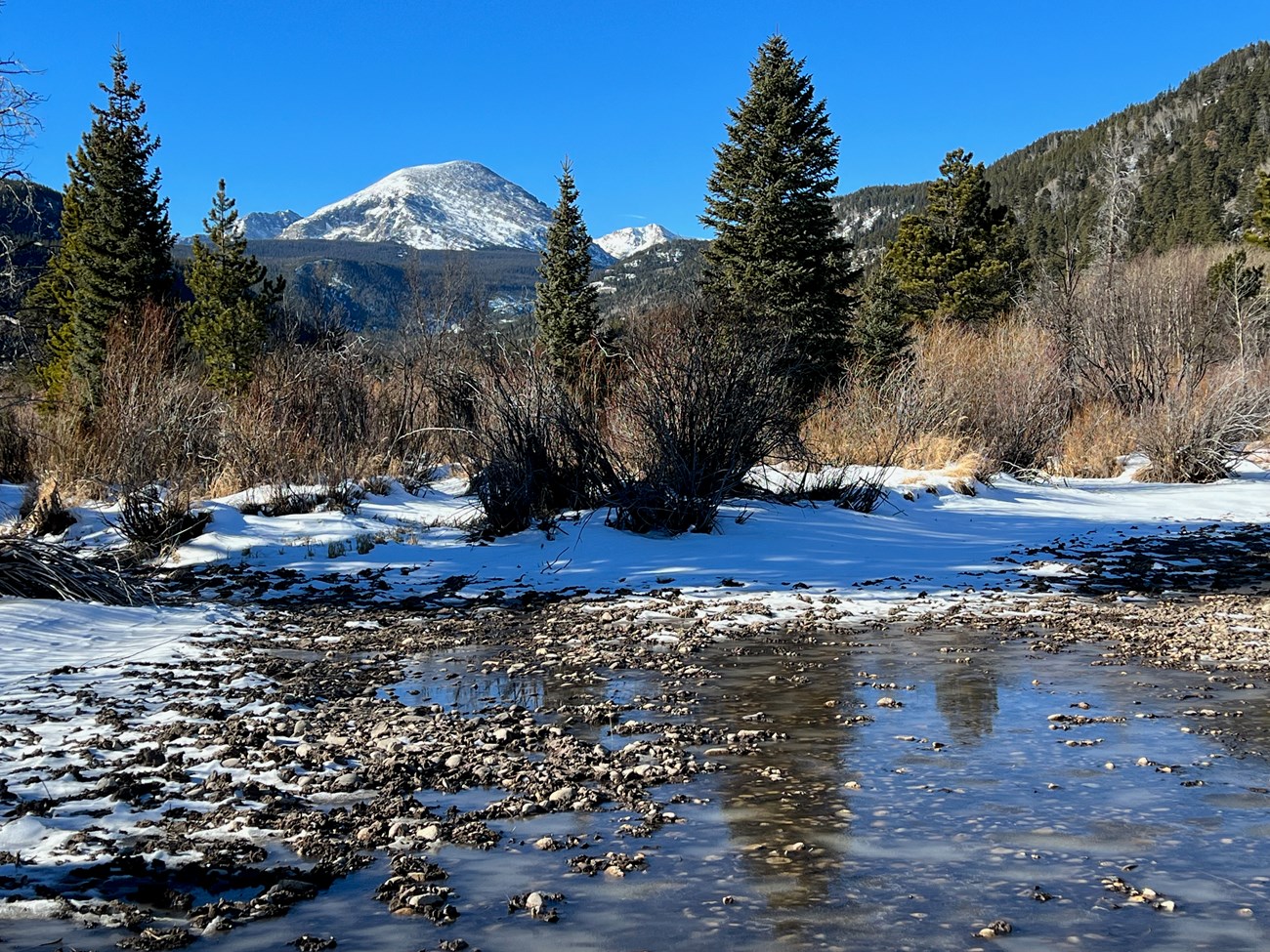 Shallow snow melting next in a gravel bed with snow covered mountain in the background.