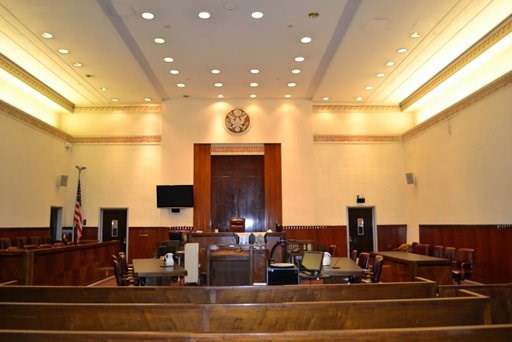 Color photo of courtroom viewed from benches for the public. A judge’s bench is against the back wall. Tables for the defendants and plaintiffs face a podium at the center. A row of seats flanks either side of the room.