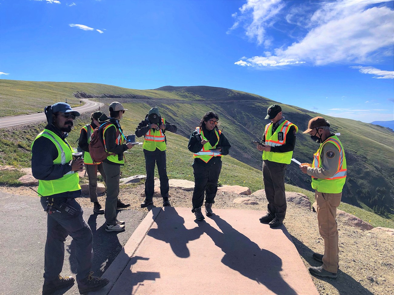 A group of people in safety vests stand in a circle talking against an alpine backdrop