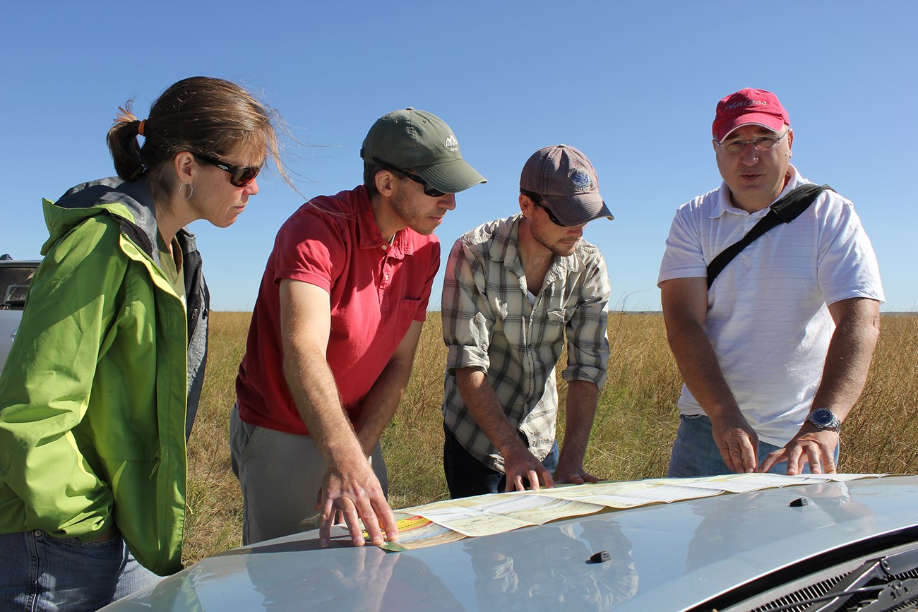 Three men and a woman look at a chart resting on the hood of a car. The older man on the right is explaining something to the others. Behind them are a large grassy field and blue skies.
