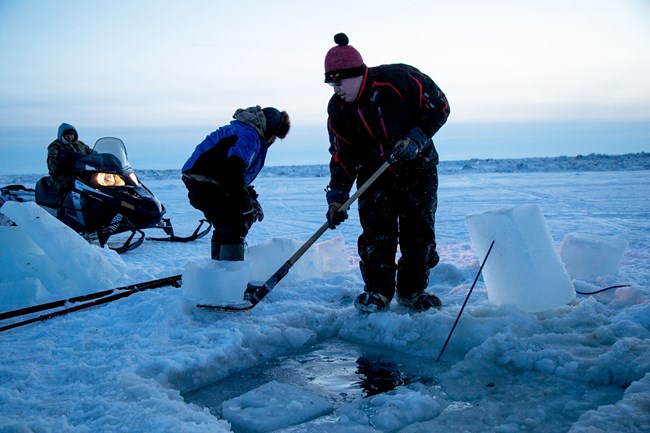 Two people work on the sea ice carving out a hole for their crab pot.
