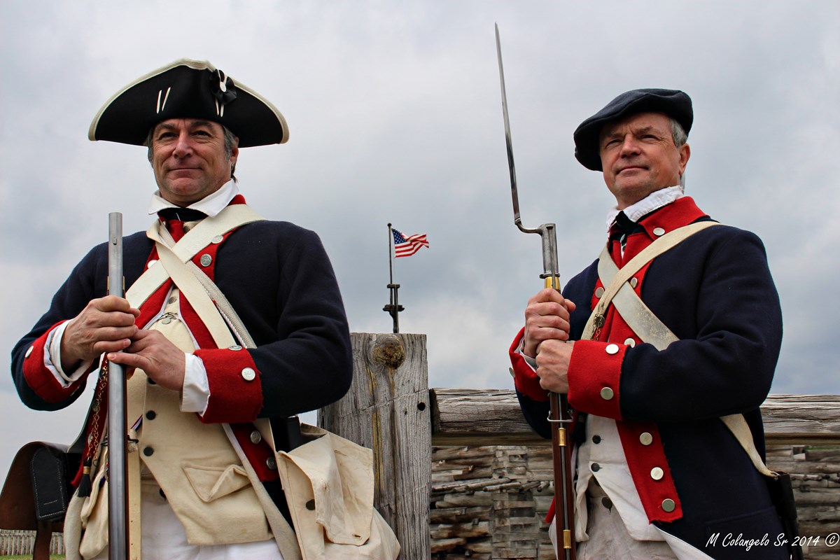 Two Continental Soldiers stand side-by-side, holding their muskets with bayonets. Between their shoulders, you can see the American flag.