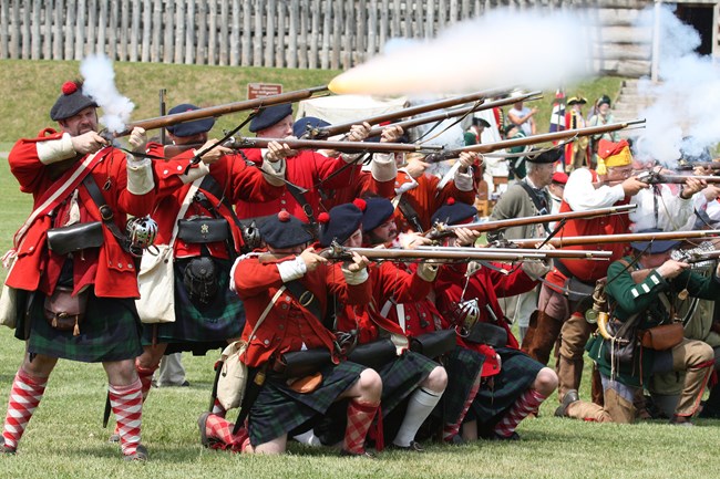 A group of men firing muskets. Two rows are in formation. One stands, the other kneels.