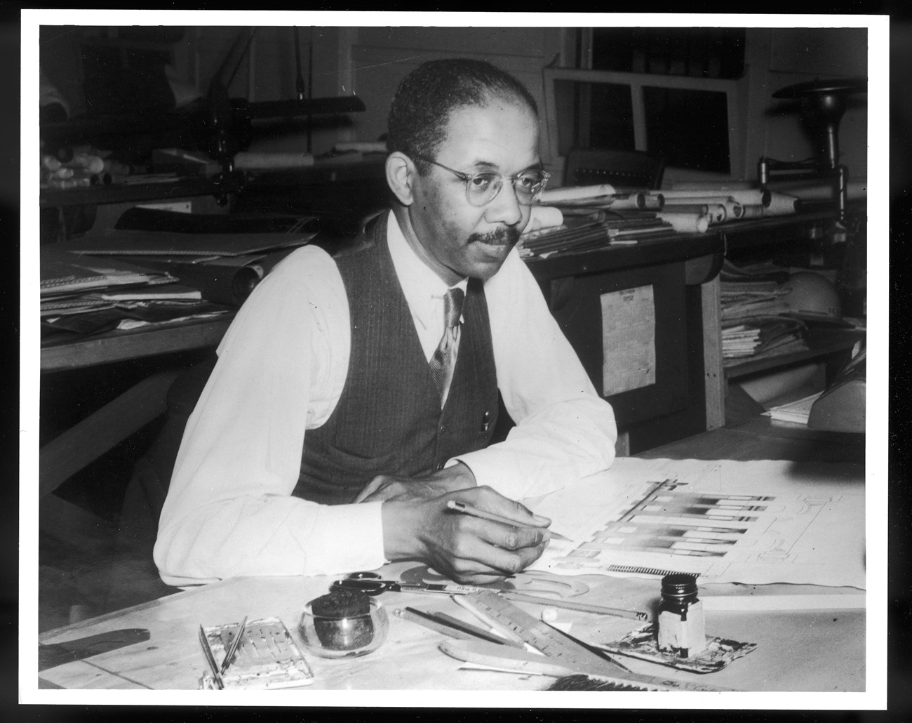 Illustrator and Arist Allan Crite sitting at his Navy Yard Desk with pen to paper.