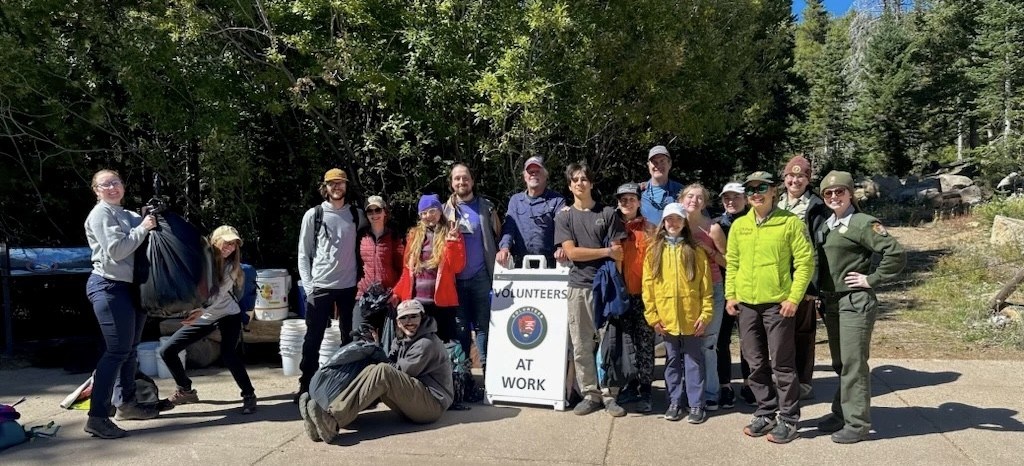 Outside at Rocky Mountain National Park, a group of around 16 volunteers and park staff are smiling next to a “Volunteers at Work” sign at a trash cleanup with trees and the sky in the background.