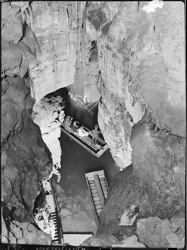 Looking down on a small wooden boat that contains several people floating in a small body of water inside a rocky cave.