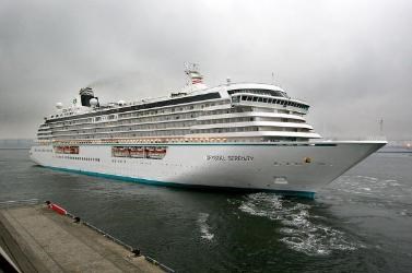 A large multi-story ship sits in the ocean near a dock in preparation for a cruise.
