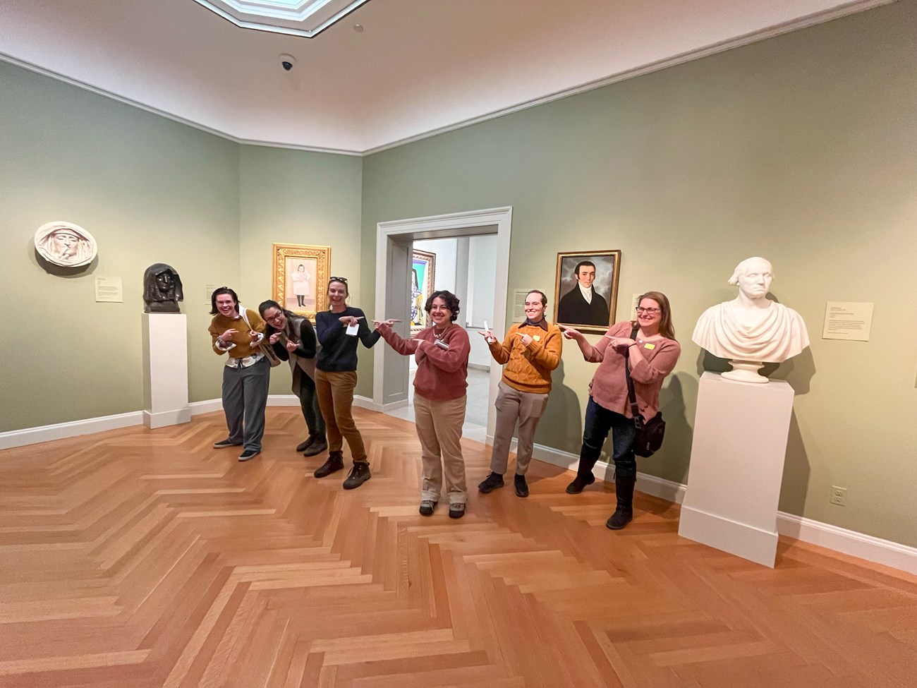 MABI Curatorial 2024 six people stand in art gallery museum exhibit pointing and each other and smiling