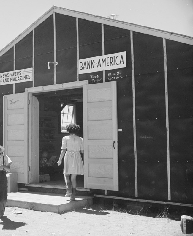 Black and white photo of a Japanese woman in a white dress entering a camp building. To the left of the door a sign reads “Newspapers and Magazines”; to the right, a sign reads “Bank of America.”