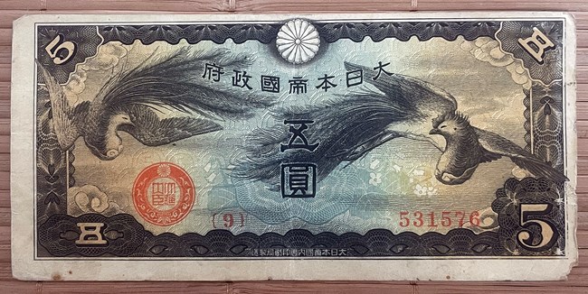 Printed bank note. The border is black with a white “5” in the upper left and lower right corners. Two birds printed in black fly through the green center of the design. A red stamp in the lower left, and a red serial number in the lower right.