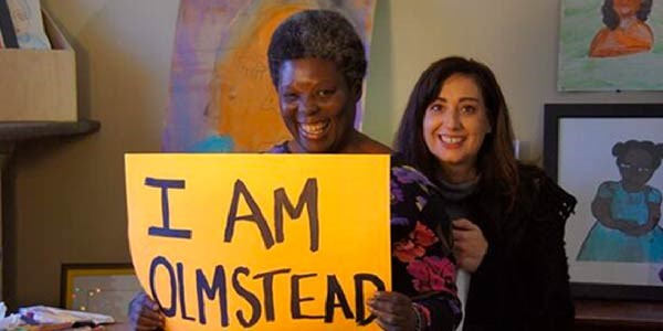 Two women standing in an art studio with hand drawn images in frames on the wall behind them. One woman is holding a sign that reads "I Am Olmstead".